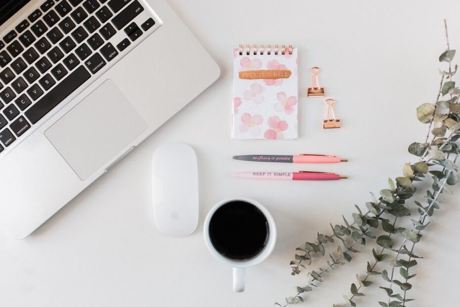 Minimalist writing workspace for a beginning writer with laptop, coffee, pink pens, floral ‘Keep It Simple’ notepad, and greenery on a white desk.