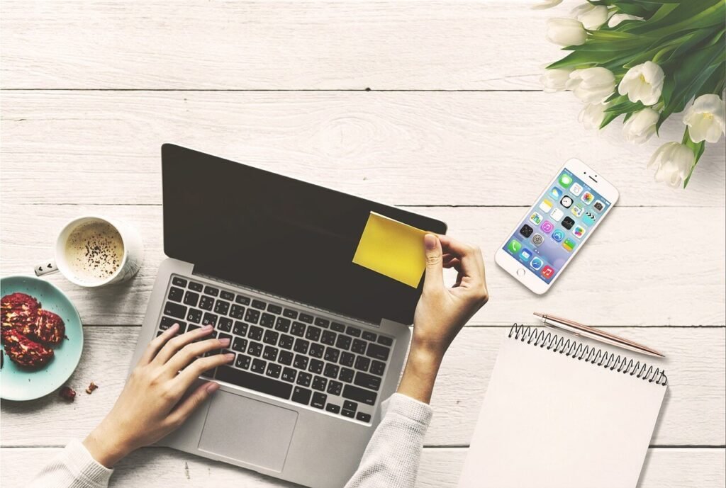 laptop with hands on keyboard and putting post it note on it and with coffee cup, cookies, notebook, pencil, tulips on a white burlap desk from above.